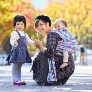 縦ストライプのベビーラップで赤ちゃんをおんぶするお母さんと手をつなぐ子ども