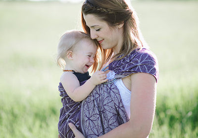 麦畑の中で女の子を紫色のスリングで抱っこするお母さん