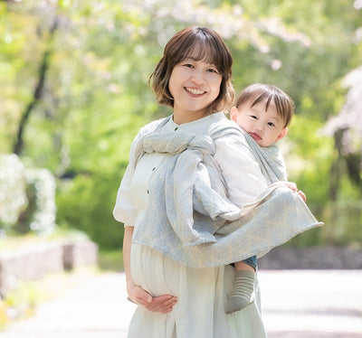 妊娠後期のママが淡いブルーの葉っぱ模様のベビーラップで上の子どもを嬉しくおんぶしている