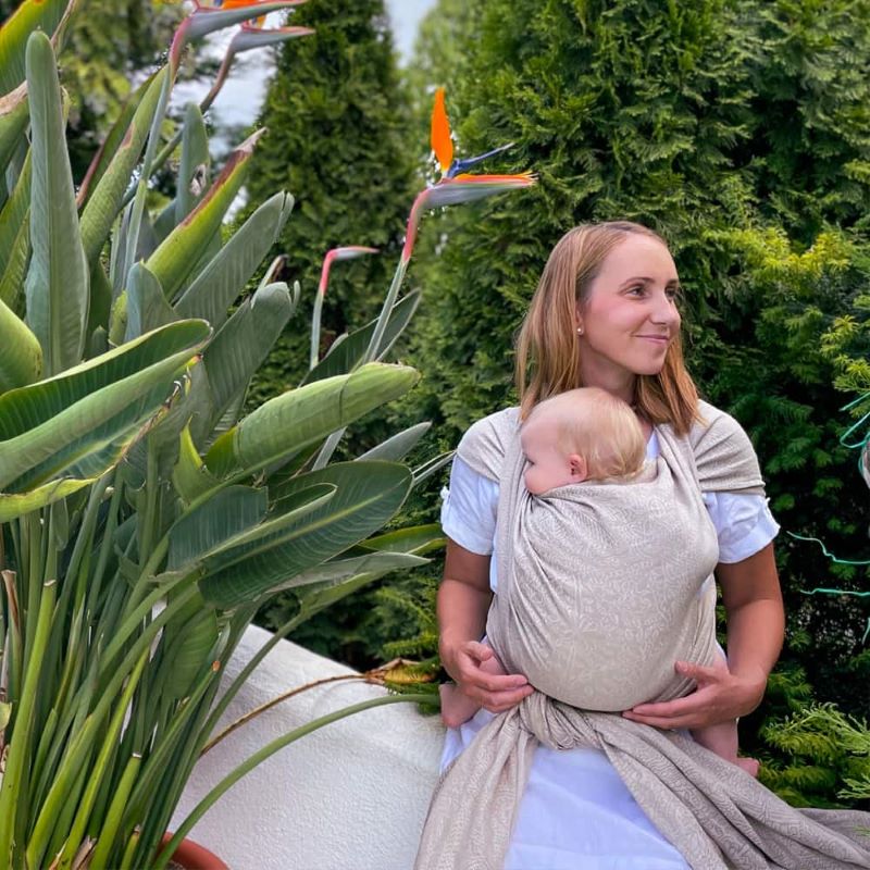 ベージュのベビーラップで赤ちゃんを抱っこしている女性が庭を散歩している