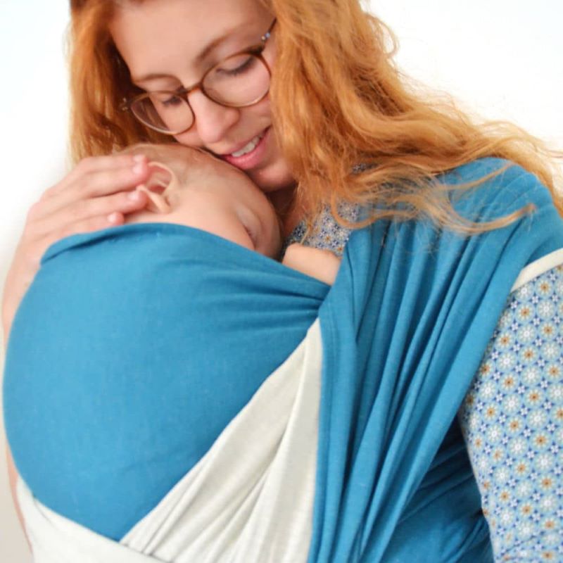 青いストレッチラップで眠る赤ちゃんを抱っこして幸せそうに微笑む女性