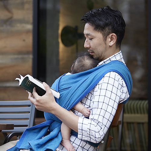 青いベビーラップで赤ちゃんを抱き、ベンチに座って本を読むパパ