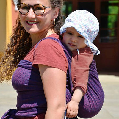 紫のベビーラップで赤ちゃんをおんぶする眼鏡をかけた白人女性